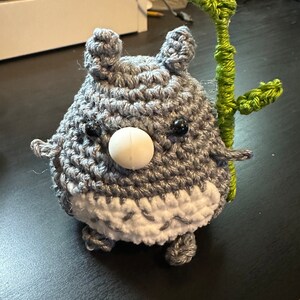 May include: A gray crocheted stuffed animal with a white nose and black eyes, holding a green crocheted stick with leaves.