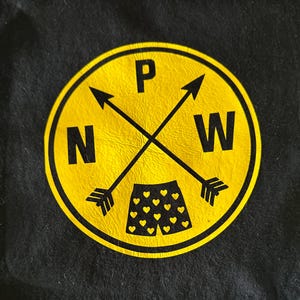 May include: A black t-shirt featuring a circular yellow graphic. The design includes two crossed black arrows, the letters N, P, and W, and a pair of black shorts patterned with small yellow hearts centered at the bottom of the circle.