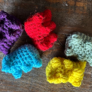 May include: A collection of six small, handmade crochet items in various colors: purple, red, blue, green, and yellow. The items are arranged on a wooden surface, showcasing their textured, three-dimensional design.