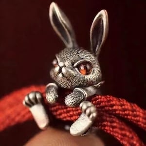 Pode incluir: Anel detalhado de coelho prateado com olhos vermelhos e expressão severa. O coelho segura um cordão vermelho, mostrando detalhes intrincados. O anel é uma peça de joalharia única.