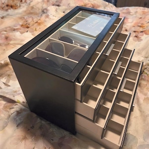 May include: A black sunglasses organizer with a glass top and multiple pull-out drawers. The top compartment holds several pairs of sunglasses. The drawers are divided into small compartments. The organizer sits on a floral patterned bedspread.