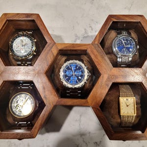 May include: A wooden watch display case with a honeycomb design, holding five wristwatches. The watches have various band and face styles, including silver, gold, and blue. The case is made of dark wood.