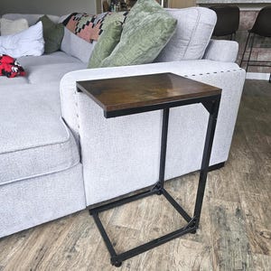 May include: A dark brown, rectangular side table with a black metal frame. The table is designed to slide over the arm of a sofa. The tabletop is made of wood and the frame is made of metal.