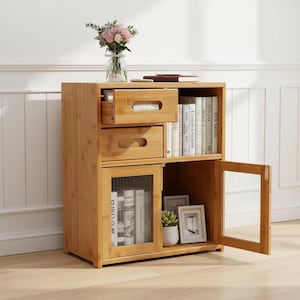 Puede incluir: Un mueble de almacenamiento de bambú marrón claro con dos cajones, una puerta de armario con panel de vidrio y estanterías abiertas. El mueble contiene libros, objetos decorativos y una pequeña planta. Un jarrón de flores rosas encima.