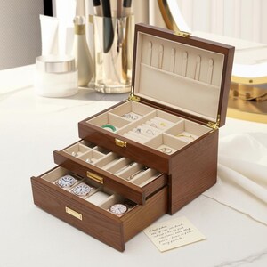 May include: A dark wooden jewelry box with multiple compartments and drawers, open to display jewelry. Rings, bracelets, and watches are visible. The interior is lined with a light-coloured fabric. The box has a gold-coloured clasp and is made of dark wood.