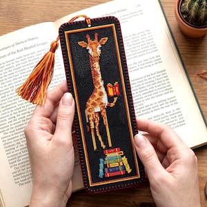 Puede incluir: Un marcapáginas rectangular negro de punto de cruz con una jirafa que sostiene un libro naranja abierto, de pie sobre una pila de libros coloridos. El marcapáginas tiene una borla decorativa naranja y amarilla y un borde cosido.
