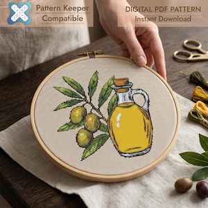 May include: A cross-stitch embroidery project in a round wooden hoop featuring a design of a glass cruet filled with yellow olive oil next to a branch with green olives and leaves. The text reads Pattern Keeper Compatible and Digital PDF Pattern.