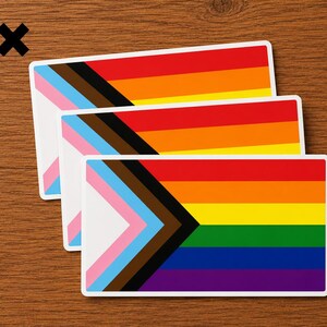 May include: Three rectangular stickers featuring the Progress Pride flag design. Each sticker displays horizontal stripes of red, orange, yellow, green, blue, and violet, with a chevron on the left side containing white, pink, light blue, brown, and black.