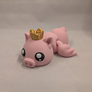 May include: A pink pig figurine wearing a gold crown. The pig has large, round eyes and a small snout. The figurine is lying down, with its legs outstretched. The overall design is cartoonish.