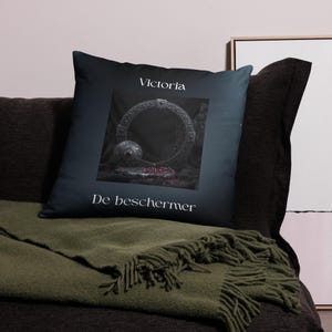 Può includere: Cuscino decorativo con sfondo blu scuro con un ornato disegno circolare color argento con una gemma rossa. Il cuscino presenta il testo "Victoria" e "De beschermer" in bianco.