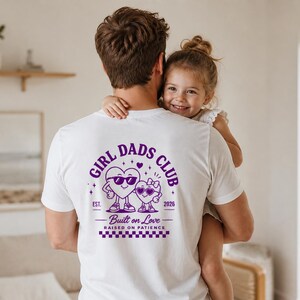 Peut inclure: T-shirt blanc avec un motif violet « GIRL DADS CLUB » avec deux personnages en forme de cœur portant des lunettes de soleil. Le texte « Built on Love, Raised on Patience » est en dessous. Un homme porte une jeune fille.