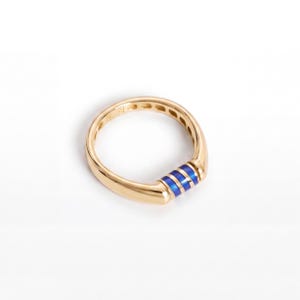May include: A gold ring featuring a smooth, polished band with a central decorative section. This section consists of three vibrant blue enamel stripes separated by thin gold lines, creating a distinct contrast against the gold metal.