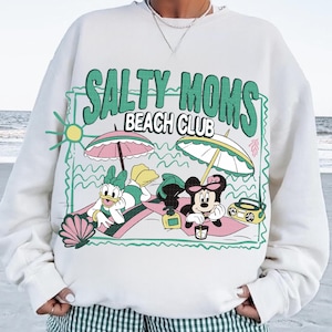 May include: White sweatshirt with a graphic featuring Daisy Duck and Minnie Mouse relaxing at the beach under umbrellas. The text "SALTY MOMS BEACH CLUB" is in green. A seashell, sun, and radio are also included.