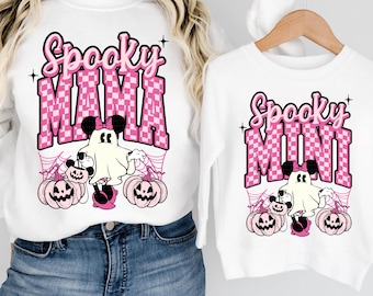 May include: White sweatshirts with Halloween-themed graphics. One says "Spooky MAMA" and the other says "Spooky MINI" in pink checkered letters. Both shirts feature a ghost, pumpkins, and spiderwebs.