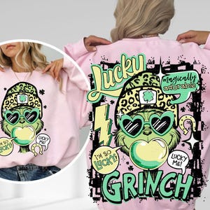 K&ouml;nnte beinhalten: Ein hellrosa Sweatshirt mit einem Cartoon-Grinch-Design. Der Grinch tr&auml;gt herzf&ouml;rmige Sonnenbrillen und einen Leopardenhut. Das Design enth&auml;lt die W&ouml;rter "Lucky" und "Grinch" in Gr&uuml;n und Schwarz, mit Textblasen mit der Aufschrift "I'm so lucky!" und "Lucky me!"