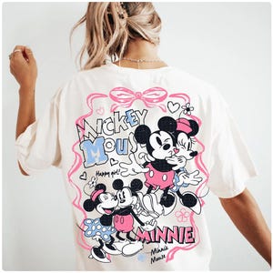 May include: Cream-colored t-shirt featuring a graphic of Mickey and Minnie Mouse embracing. The design includes the words "Mickey Mouse" and "Minnie" in pink and blue, with hearts and floral accents. The shirt has short sleeves.