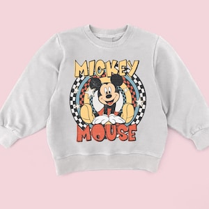 May include: A light grey sweatshirt with a retro Mickey Mouse graphic. The design features the words "Mickey Mouse" in yellow and red, with Mickey in the centre. The graphic is set against a checkered and star-filled background.
