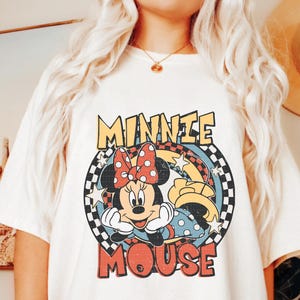 Puede incluir: Camiseta color crema con un dise&ntilde;o retro de Minnie Mouse. El dise&ntilde;o incluye la cara de Minnie, un lazo de lunares y las palabras "MINNIE MOUSE" en una fuente vintage. El gr&aacute;fico est&aacute; sobre un fondo a cuadros y lleno de estrellas.