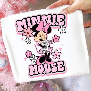May include: White fabric featuring a Minnie Mouse graphic. The design includes Minnie Mouse in a pink dress, holding a small gray bunny, surrounded by pink and white flowers, and the words "MINNIE MOUSE" in pink bubble letters.
