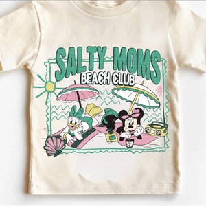 May include: Cream-colored t-shirt featuring the text "SALTY MOMS BEACH CLUB" in green. The graphic includes cartoon characters Daisy Duck and Minnie Mouse relaxing on the beach under umbrellas, with a boombox and seashell.