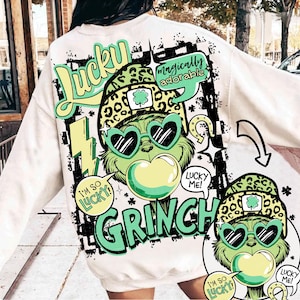 K&ouml;nnte beinhalten: Cremefarbenes Sweatshirt mit einer Grafik eines gr&uuml;nen Grinch-Charakters mit herzf&ouml;rmiger Sonnenbrille und Leopardenmuster-Hut. Text enth&auml;lt "Lucky", "Grinch" und Sprechblasen mit "I'm so lucky!" und "Lucky me!"
