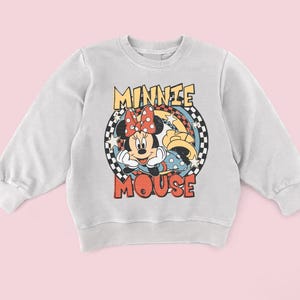 Puede incluir: Sudadera gris claro con un dise&ntilde;o de Minnie Mouse. El dise&ntilde;o incluye la cara de Minnie, un lazo rojo con lunares blancos y las palabras "MINNIE MOUSE" en una fuente retro. El dise&ntilde;o est&aacute; sobre un fondo a cuadros.