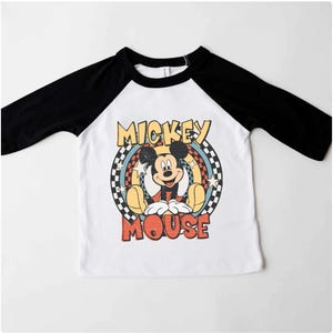 May include: A white and black raglan-style shirt with a Mickey Mouse graphic. The graphic features the words "Mickey Mouse" in a retro font. The shirt has black sleeves and a white body.