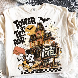 Può includere: Una t-shirt color crema con una stampa grafica di un hotel infestato, luna piena, pipistrelli, fantasmi e un topo dei cartoni animati che tiene una valigia. Il testo recita TOWER OF TERROR e HAUNTED HOTEL in un carattere stile vintage.