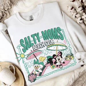 May include: White sweatshirt with a graphic that reads "SALTY MOMS BEACH CLUB" in green and pink. The graphic features cartoon characters under beach umbrellas, with a boombox and seashell.