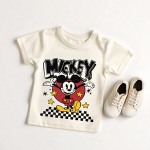 Puede incluir: Camiseta color crema con un dise&ntilde;o vintage de Mickey Mouse. El dise&ntilde;o incluye la palabra "MICKEY" en negro, Mickey Mouse en un c&iacute;rculo rojo y estrellas amarillas. Un par de zapatillas de lona color crema est&aacute;n junto a la camiseta.