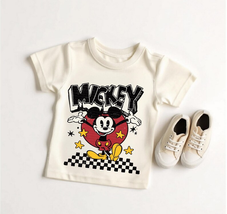 Puede incluir: Camiseta color crema con un dise&ntilde;o vintage de Mickey Mouse. El dise&ntilde;o incluye la palabra "MICKEY" en negro, Mickey Mouse en un c&iacute;rculo rojo y estrellas amarillas. Un par de zapatillas de lona color crema est&aacute;n junto a la camiseta.