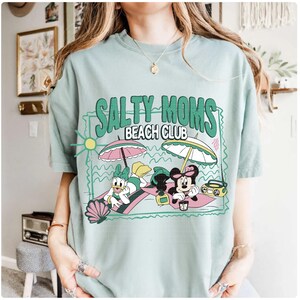 May include: A light green t-shirt with the text "SALTY MOMS BEACH CLUB" and a graphic of Daisy Duck and Minnie Mouse relaxing on the beach under umbrellas. The shirt has a relaxed fit.
