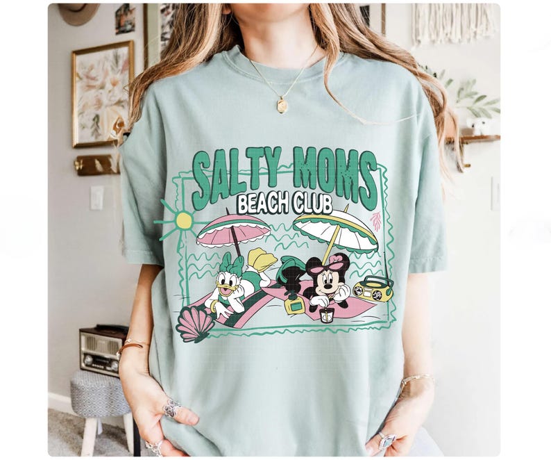 May include: A light green t-shirt with the text "SALTY MOMS BEACH CLUB" and a graphic of Daisy Duck and Minnie Mouse relaxing on the beach under umbrellas. The shirt has a relaxed fit.