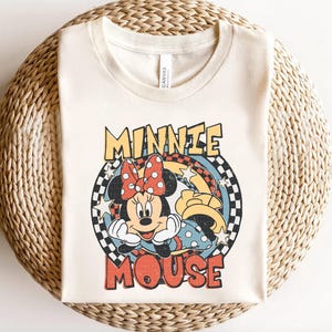 Puede incluir: Camiseta color crema con un gr&aacute;fico retro de Minnie Mouse. El dise&ntilde;o incluye la cara de Minnie, un lazo rojo con lunares y las palabras "MINNIE MOUSE" en una fuente vintage. La camiseta est&aacute; hecha de un material suave.