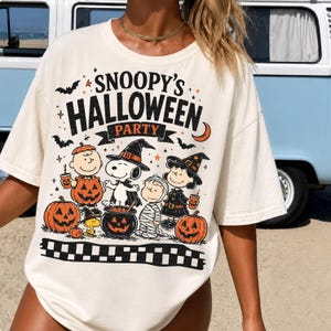 May include: An off-white short-sleeve t-shirt featuring a graphic print that reads SNOOPY'S HALLOWEEN PARTY. The design includes illustrations of Snoopy, Charlie Brown, and other characters in costumes, surrounded by pumpkins, bats, and stars.