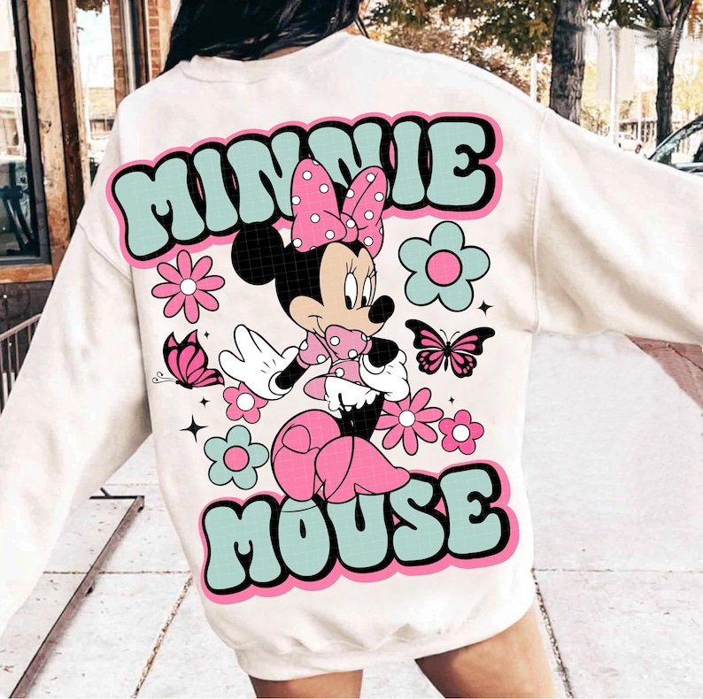 May include: Cream-colored sweatshirt featuring a colorful Minnie Mouse graphic. The design includes the words "MINNIE MOUSE" in a retro font, along with flowers and butterflies. Minnie Mouse is depicted in a pink polka-dot bow and dress.