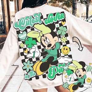 Könnte beinhalten: Cremefarbenes Sweatshirt mit einer Grafik von Minnie Mouse in Grün, die ein vierblättriges Kleeblatt hält. Das Design enthält den Text "Cutest Clover in the Park" mit einem karierten Hintergrund, Kleeblättern und Smiley-Gesichtern.