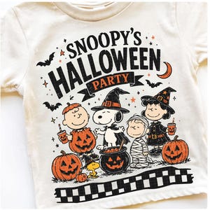May include: A cream-colored t-shirt featuring a graphic print with the text SNOOPY'S HALLOWEEN PARTY. The design includes Snoopy, Charlie Brown, Lucy, and Woodstock in costumes, surrounded by pumpkins, bats, a crescent moon, and stars.