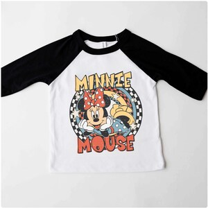 Puede incluir: Camiseta de estilo ragl&aacute;n blanca y negra con un dise&ntilde;o de Minnie Mouse, lazo rojo, estrellas y la frase "Minnie Mouse". El dise&ntilde;o est&aacute; enmarcado por un c&iacute;rculo a cuadros. Mangas negras.