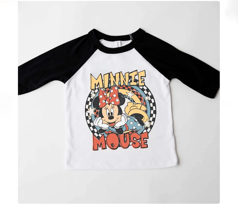 Puede incluir: Camiseta de estilo ragl&aacute;n blanca y negra con un dise&ntilde;o de Minnie Mouse, lazo rojo, estrellas y la frase "Minnie Mouse". El dise&ntilde;o est&aacute; enmarcado por un c&iacute;rculo a cuadros. Mangas negras.