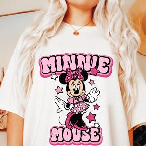 Peut inclure: T-shirt blanc avec un motif r&eacute;tro de Minnie Mouse. Le graphisme comprend les mots "MINNIE MOUSE" en lettres roses &agrave; bulles, avec Minnie Mouse au centre, entour&eacute;e d'&eacute;toiles roses. Le design a une esth&eacute;tique vintage et cartoon.