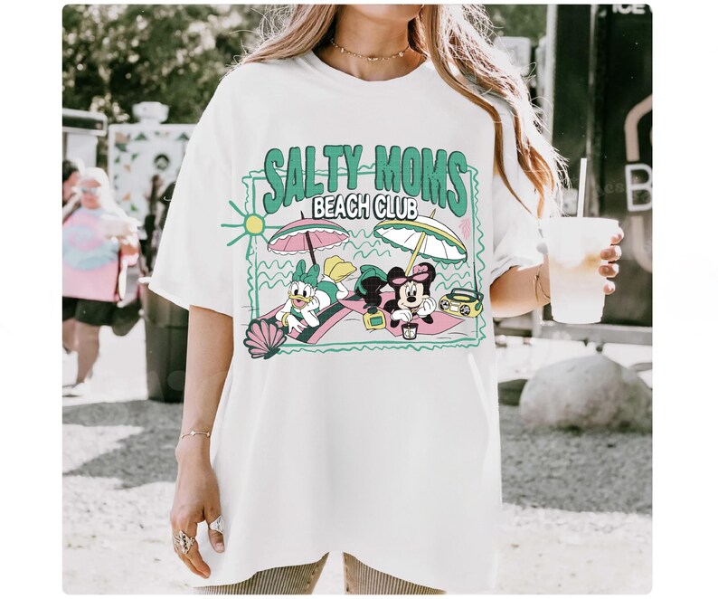 May include: White t-shirt with a graphic that reads "Salty Moms Beach Club." The graphic features cartoon characters Daisy Duck and Minnie Mouse relaxing on beach towels under umbrellas. A retro radio is also included in the design.
