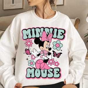 May include: White sweatshirt featuring a retro design with the text "MINNIE MOUSE" in teal and pink. The graphic includes a cartoon Minnie Mouse character, flowers, and butterflies. The design has a vintage, groovy aesthetic.