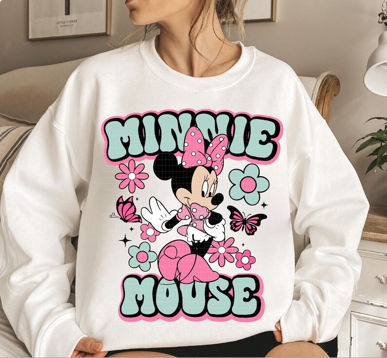 May include: White sweatshirt featuring a retro design with the text "MINNIE MOUSE" in teal and pink. The graphic includes a cartoon Minnie Mouse character, flowers, and butterflies. The design has a vintage, groovy aesthetic.