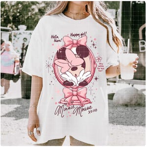 May include: White t-shirt with a Minnie Mouse graphic in a pink mirror frame and bows. The design includes the text "Hello, there! Happy girl! Minnie Mouse xoxo" and "Why settle for less fun!" A playful, whimsical design.