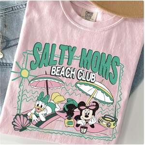 May include: Pink t-shirt with the text "SALTY MOMS BEACH CLUB" in green. The graphic features cartoon characters relaxing on the beach under umbrellas. A seashell, sun, and boombox are also included in the design.