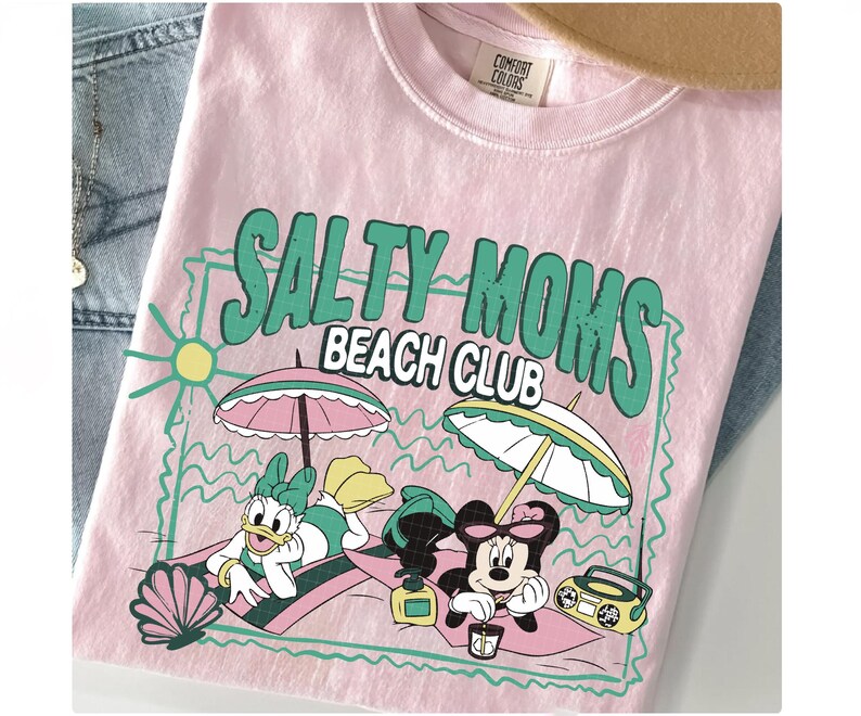 May include: Pink t-shirt with the text "SALTY MOMS BEACH CLUB" in green. The graphic features cartoon characters relaxing on the beach under umbrellas. A seashell, sun, and boombox are also included in the design.