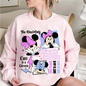 May include: A light pink sweatshirt featuring three Minnie Mouse graphics with bows. The text includes "The One & Only", "Minnie Mouse", and "Cute is a Lifestyle". The design also includes the text "Sweet as Can Be".