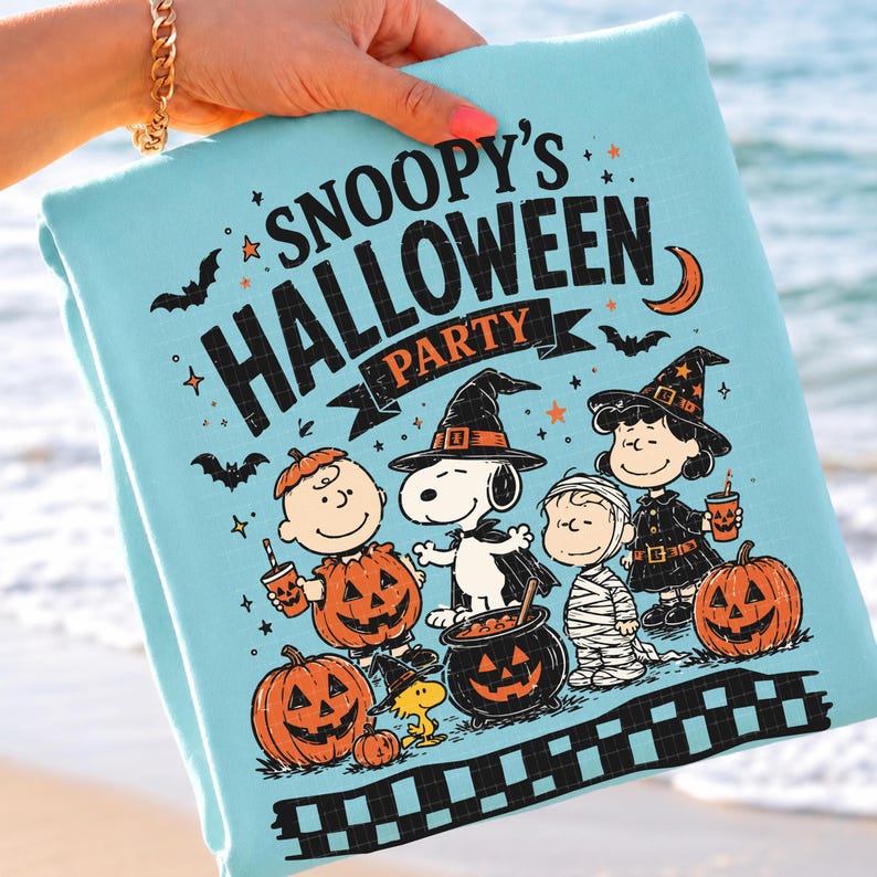 May include: A light blue t-shirt featuring a graphic print with the text SNOOPY'S HALLOWEEN PARTY. The design includes Snoopy, Charlie Brown, and other characters in costumes, surrounded by jack-o'-lanterns, bats, stars, and a crescent moon.