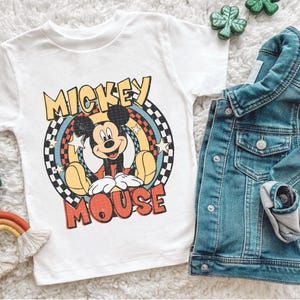 May include: White t-shirt with a Mickey Mouse graphic and the words "Mickey Mouse". A denim jacket is folded nearby. Small rainbow and shamrock decorations are also visible.